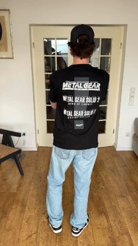 Persona che indossa una T-shirt nera di Metal Gear Solid, jeans azzurri, cappellino nero e sneakers, in piedi al chiuso di fronte a una porta con pannelli di vetro.
