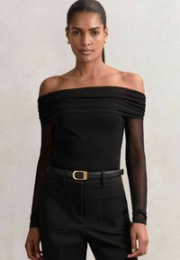 Body negru, cu umerii descoperiți, cu detalii plisate și mâneci lungi din material translucid, asociat cu pantaloni negri cu talie înaltă și o curea cu cataramă aurie.