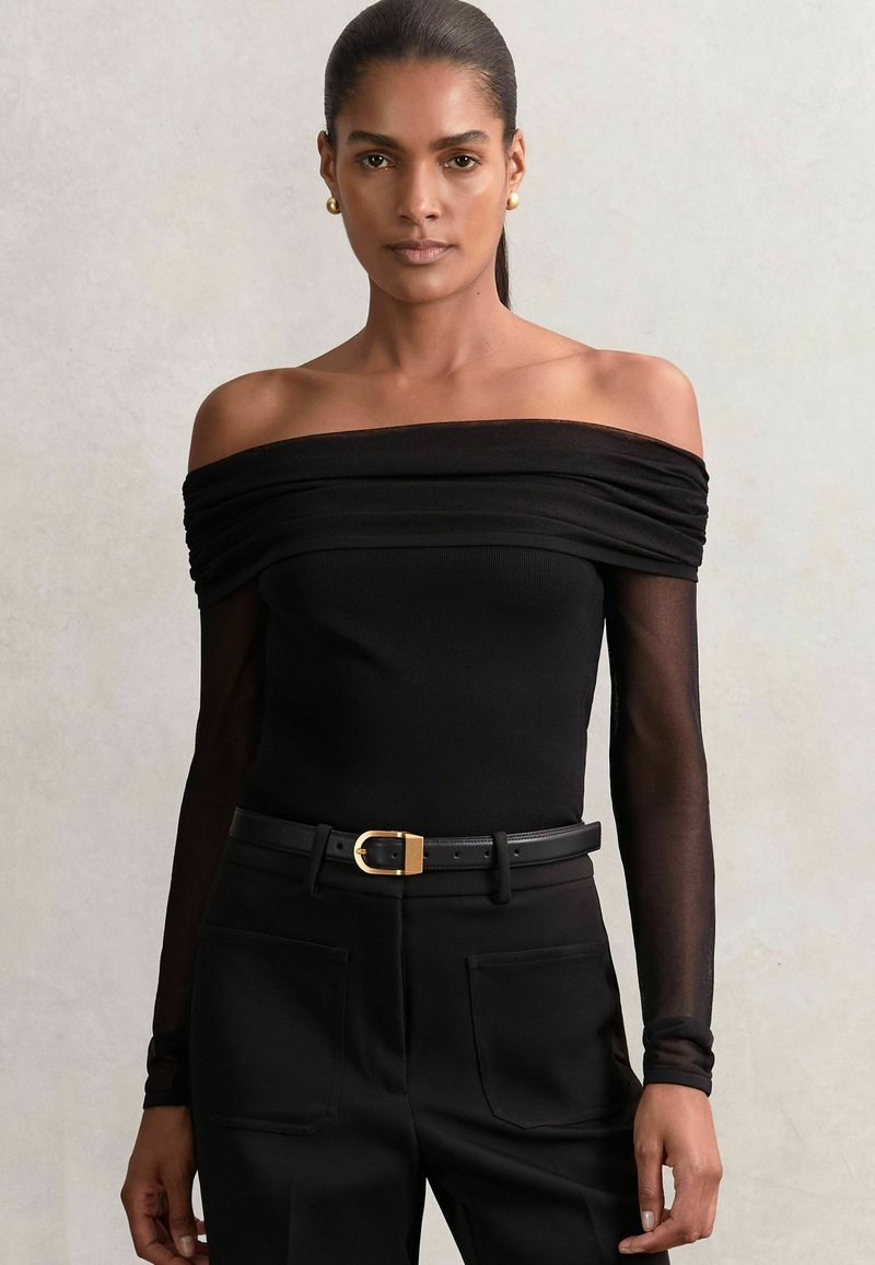 Body negru, cu umerii descoperiți, cu detalii plisate și mâneci lungi din material translucid, asociat cu pantaloni negri cu talie înaltă și o curea cu cataramă aurie.