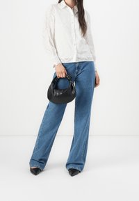 Kvinna i vit broderad blus och vida blå jeans som håller en liten svart läderhandväska, stående på vit bakgrund.