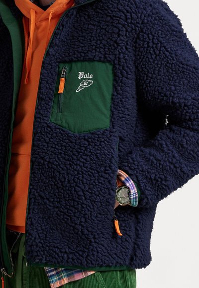 Marineblå fleecejakke med grøn lomme med lynlås på brystet, iført over en orange hoodie og lagdelt med ternede manchetter og grønne kordfløjlbukser.