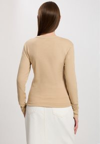 Beige Langarmoberteil mit Rundhalsausschnitt, aus weichem Material, kombiniert mit einem weißen Rock mit aufgesetzten Taschen und sichtbaren Nähten.