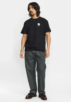 Hombre con cabello oscuro hasta los hombros, lleva una camiseta negra con un pequeño logo blanco, pantalones gris oscuro y zapatos marrón oscuro, de pie frente a un fondo blanco.