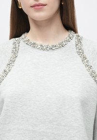 Sweatshirt cinza com decote bordado com contas transparentes e pérolas ao longo dos ombros, com textura suave e macia.