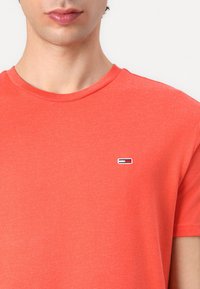Tricou roșu coraliu din bumbac moale. Dispune de un guler rotund și un mic logo brodat pe pieptul stâng. Design simplu, cu mâneci scurte.