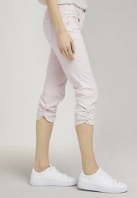 Jasnoróżowe skórzane spodnie 3/4 z marszczeniami na zakończeniu, wykonane z materiału dżinsowego, w połączeniu z białymi trampkami na neutralnym tle.