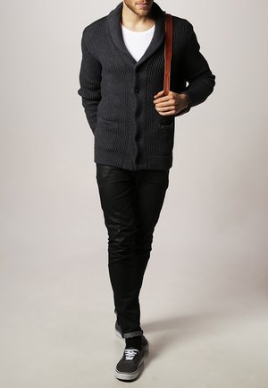 Cárdigan de punto gris oscuro con cuello de chal, que cuenta con botones y dos bolsillos frontales. Usado sobre una camisa blanca y combinado con pantalones negros y zapatillas.
