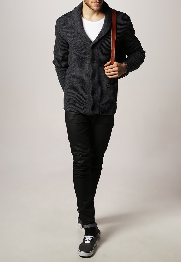 Mörkgrå stickad cardigan med sjalkrage, med knappar och två framfickor. Bärs över en vit skjorta och kombineras med svarta byxor och sneakers.