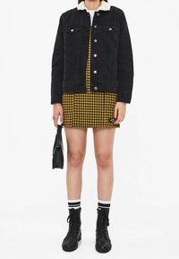 Chaqueta de mezclilla negra con cuello de borreguito, vestido a cuadros amarillos, botas de combate negras y calcetines a rayas. Sosteniendo un pequeño bolso negro.