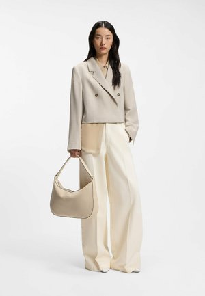Woman in beige cropped blazer, wide-leg cream trousers, and beige handbag standing against white background.