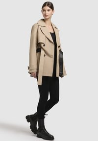 Beige trenchcoat met dubbele knopen en een zwarte riem, gecombineerd met zwarte leggings en chunky zwarte laarzen. Model staat voor een effen achtergrond.