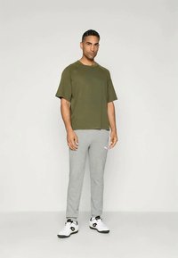 Homem de pé contra um fundo branco, vestindo uma camisa de manga curta verde oliva, calças de treino cinza claro e ténis brancos.