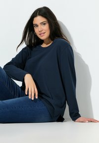 Navy-blaues Langarmoberteil aus weichem Material mit lockerer Passform, runder Ausschnitt und seitlichen Schlitzen, kombiniert mit dunkelblauen Jeans auf einem weißen Hintergrund.
