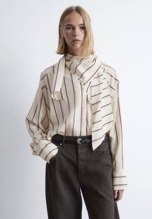 Eine blonde Person trägt eine cremefarbene Bluse mit vertikalen Streifen und einem angenähten Schal, kombiniert mit dunklen Hosen und einem schwarzen Gürtel.