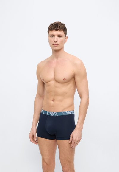 Άντρας χωρίς πουκάμισο όρθιος, φοράει σκούρα μπλε ανδρικά εσώρουχα boxer με ζώνη μέσης με λογότυπο, σε απλό φωτεινό φόντο.