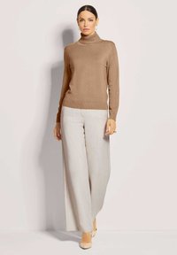 Brauner Rollkragenpullover aus weichem Stoff, tailliert. Kombiniert mit hellgrauen, weiten Hosen mit dezentem Muster. Neutrale Schuhe.