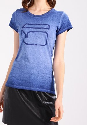 Femme portant une chemise à manches courtes bleue avec un design abstrait sombre et une jupe noire brillante, debout devant un fond uni.