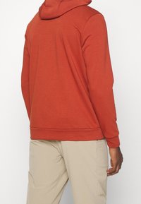Red sweatshirt with a hood, made of soft fabric. Features ribbed cuffs and hem, with stitching details. Paired with beige trousers.