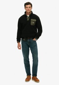 Sort fleece-pullover med en grøn brystlomme, lagdelt over en grøn skjorte, parret med blå jeans og brune snørebåndsstøvler.