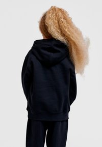 Svart luvtröja med avslappnad passform, utrustad med en känguruficka och ribbade muddar, bärs tillsammans med matchande byxor. Texturerat lockigt hår syns.