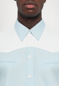 Camicia azzurro chiaro con colletto appuntito, yoke bianco e due piccole tasche sul petto con dettagli a freccia. Tessuto liscio.