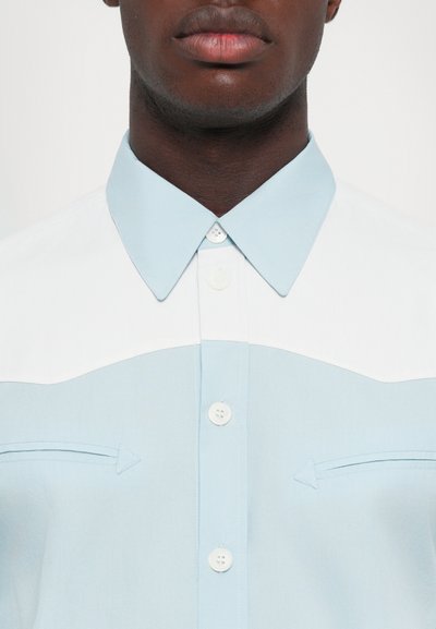 Camicia azzurro chiaro con colletto appuntito, yoke bianco e due piccole tasche sul petto con dettagli a freccia. Tessuto liscio.