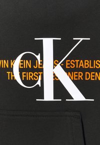 Černá mikina s velkým bílým logem "CK" a oranžovým textem "CALVIN KLEIN JEANS - ZALOŽENA PRVNÍM ZÁKLADNÍM DENIMEM." Hladká textura.