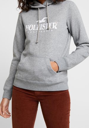 Haut à capuche gris avec une poche kangourou, un cordon de serrage à la capuche, et un logo "Hollister California" agrémenté d'accents floraux. Poignets et ourlet côtelés.