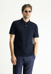 Polo shirt navy in misto cotone, con colletto classico, tre bottoni e maniche corte, abbinato a pantaloni di lino blu scuro.