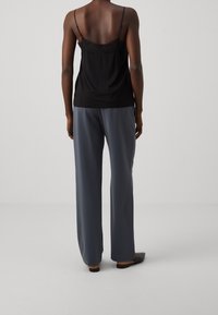 Vrouw die met haar rug naar de camera staat, gekleed in een zwarte met kant afgewerkte cami-top, losse grijze broek en zwarte instappers, voor een effen achtergrond.