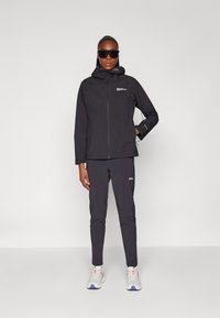 Black waterproof jacket and pants with a fitted design, featuring a hood and zippers. Worn with white running shoes and sunglasses.