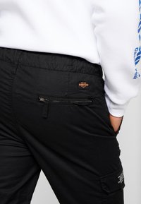 Zwarte broek met een elastische tailleband, voorzien van een ritssluiting achterzak en een klein label op de taille. De stof lijkt textuur te hebben.