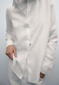 Wit linnen overhemd met knopen aan de voorkant en opgerolde manchetten, gedragen over een gestructureerde top. Zilveren hangerketting hangt om de hals.
