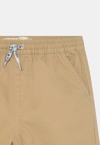 Pantaloni beige con vita elastica e cordini bianchi, con testo blu e cuciture di tasche laterali visibili sul lato destro.