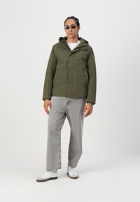 Oliven grønn vanntett jakke med hette, frontglidelås og to lommer, kombinert med lysegrå løse jeans og hvite sneakers.