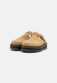 Zoccoli in suede marrone con fibbia, caratterizzati da una punta arrotondata e una suola nera spessa e texture, accentuata da cuciture gialle.