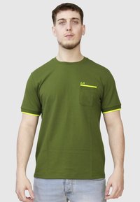 T-shirt verde oliva a maniche corte in tessuto morbido con accenti gialli sulle maniche e una tasca. Presenta un logo minimalista sul petto.