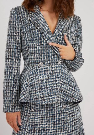 Kvinde iført en blå, hvid og sort tweed-peplumblazer med sølvknapper og matchende nederdel, hånd peger på blazeren.