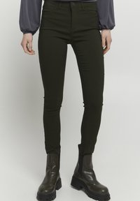 Tmavě zelené skinny džíny s předními kapsami, přiléhavý střih a mírný lesk. Nesené s masivními černými kotníkovými botami.