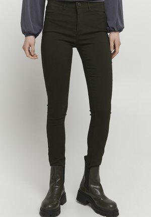 Tmavě zelené skinny džíny s předními kapsami, přiléhavý střih a mírný lesk. Nesené s masivními černými kotníkovými botami.