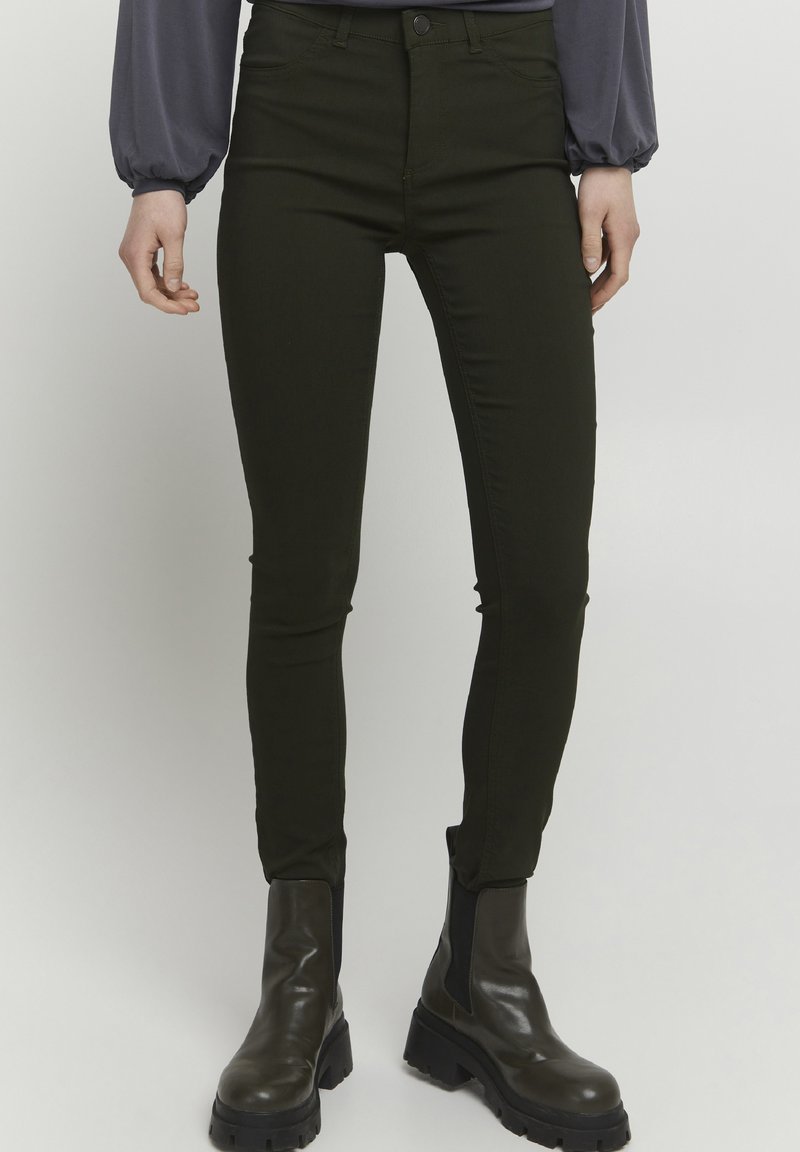 Jean skinny vert foncé avec poches avant, design ajusté et léger éclat. Porté avec des bottines noires à semelles épaisses.