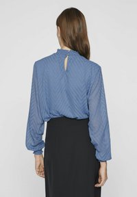 Niebieska bluzka z teksturowanym wzorem chevronu, przezroczystymi długimi rękawami, falbanką przy dekolcie oraz otworem w kształcie łezki z tyłu. Połączona z czarną spódnicą.