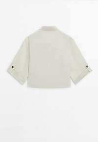 Light beige cropped shirt with a subtle texture, featuring a collar and rolled-up sleeves secured with buttons. Simple and minimalistic design.