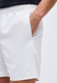 Κοντινό πλάνο ενός ατόμου που φοράει λευκά σορτς με ελαστική μέση, με το χέρι να ακουμπάει μέσα στην δεξιά τσέπη, μπροστά σε απλό φόντο.