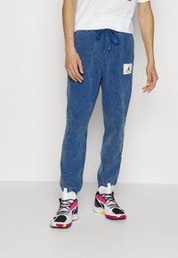 Blaue Baumwoll-Jogginghosen mit einer ausgewaschenen Textur, ausgestattet mit einem Kordelzugbund und einem Logo-Patch. Getragen mit mehrfarbigen Sportschuhen.