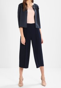 Veste marine à pois avec une taille cintrée, associée à un haut rose clair et un pantalon large marine. Des chaussures beige à bride à la cheville complètent la tenue.