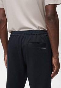 Zwarte sportieve joggers met een gestructureerde afwerking, elastische tailleband en een klein achterzakje. Kenmerkende label: "MNG PERFORMANCE."