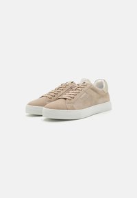 Beige semskinnsneakers med hvite gummisåler, med flate snørebånd og et minimalisk design. Ingen synlige mønstre eller dekorasjoner.