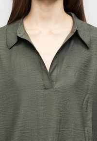 Κοντινό πλάνο ενός ατόμου που φοράει μια υφασμάτινη πουκάμισο σε ελαιώδες πράσινο με γιακά και V-σχήμα λαιμόκοψη, εμφανίζοντας τον λαιμό και το πάνω μέρος του στήθους.