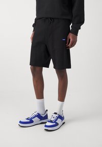 Shorts de sport noirs avec cordon de serrage ajustable, arborant un petit logo bleu "HUGO". Associés à des chaussettes blanches et des baskets avec des accents bleus.